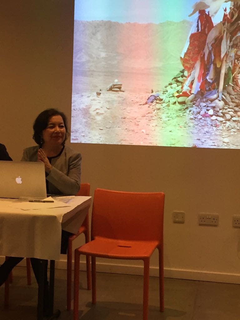 Rahima Mahmut, sitting against the backdrop of a photograph of a Sufi shrine in the Taklamakan Desert in Xinjiang.