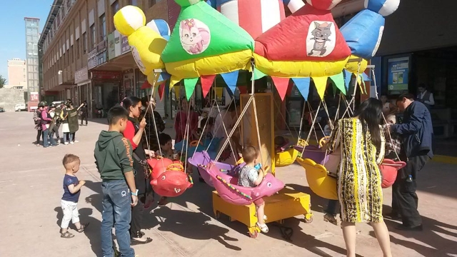 Contrasting images of Xinjiang: children playing on the carousel against a background of the Hotan market “home guards” practicing a drill.